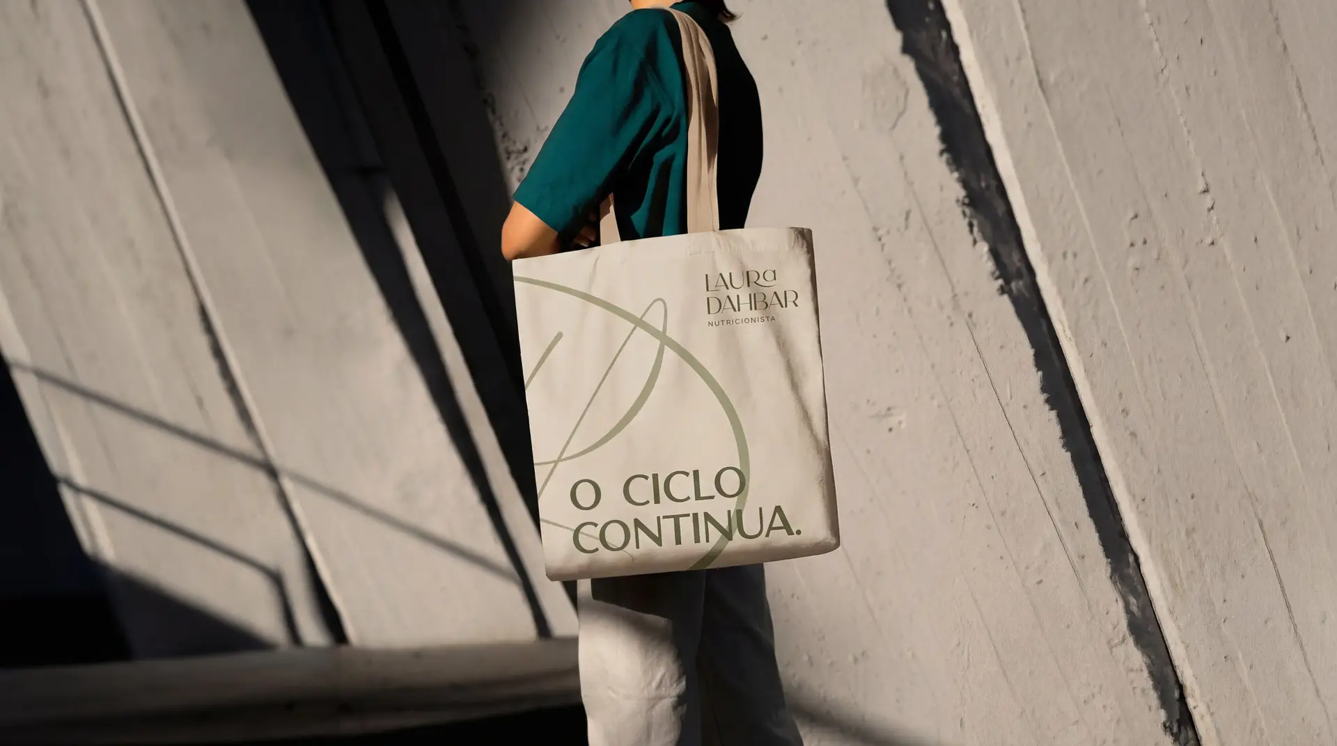 Mulher com uma ecobag creme no ombro, com o monograma LD e a frase “O ciclo continua.”, na frente de um muro de concreto com sombra.