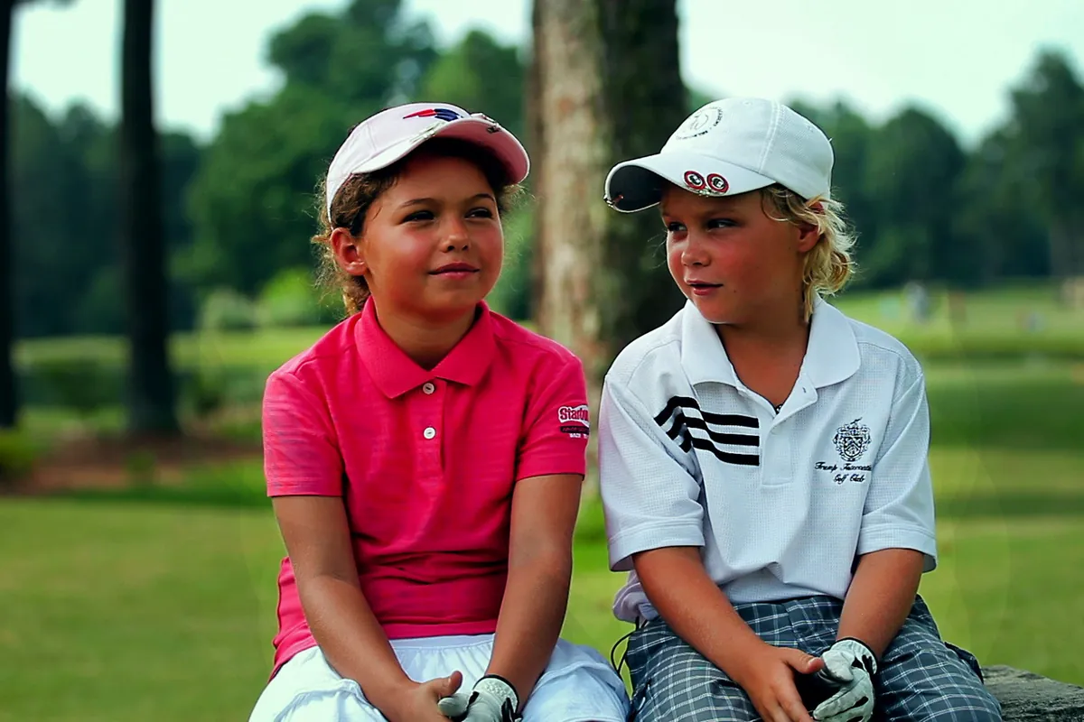 Duas crianças com boné e roupa de golfe sentadas lado a lado num campo, numa cena do documentário The Short Game